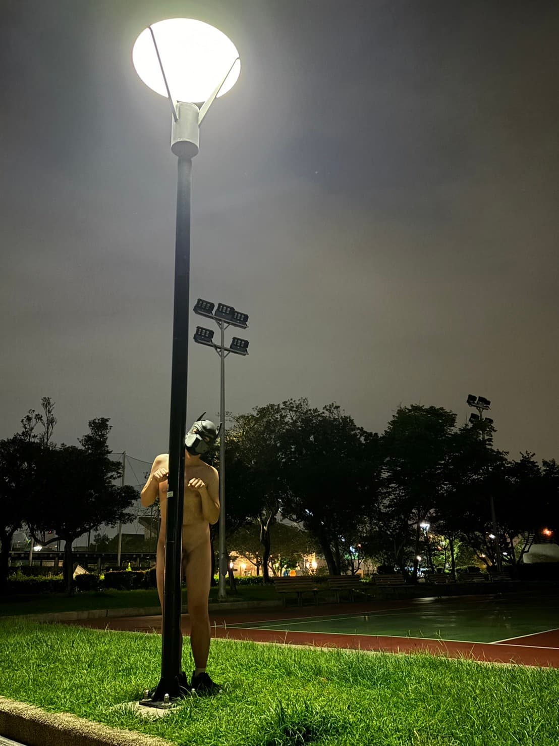 A lonely nude figure wearing a Batman-style mask stands frozen against a streetlight pole in a quiet nighttime park beside a sports court, the bright glow contrasting with dark trees and empty benches to create a surreal, timeless mood of quiet unease and artistic isolation.