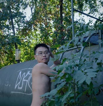 A young man with glasses leans against lush green plants beside a graffiti-covered industrial tank in a wooded area, captured in natural daylight with an intimate, contemplative mood and artistic elements of vulnerability and nature's embrace.