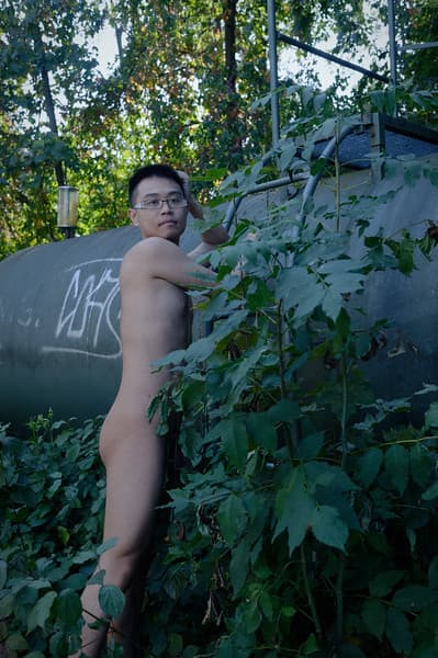 A bare young man with glasses stands in quiet contemplation, one hand resting on a large metal tank overgrown with lush green foliage and graffiti, evoking solitude and introspective calm amid the untamed forest setting.