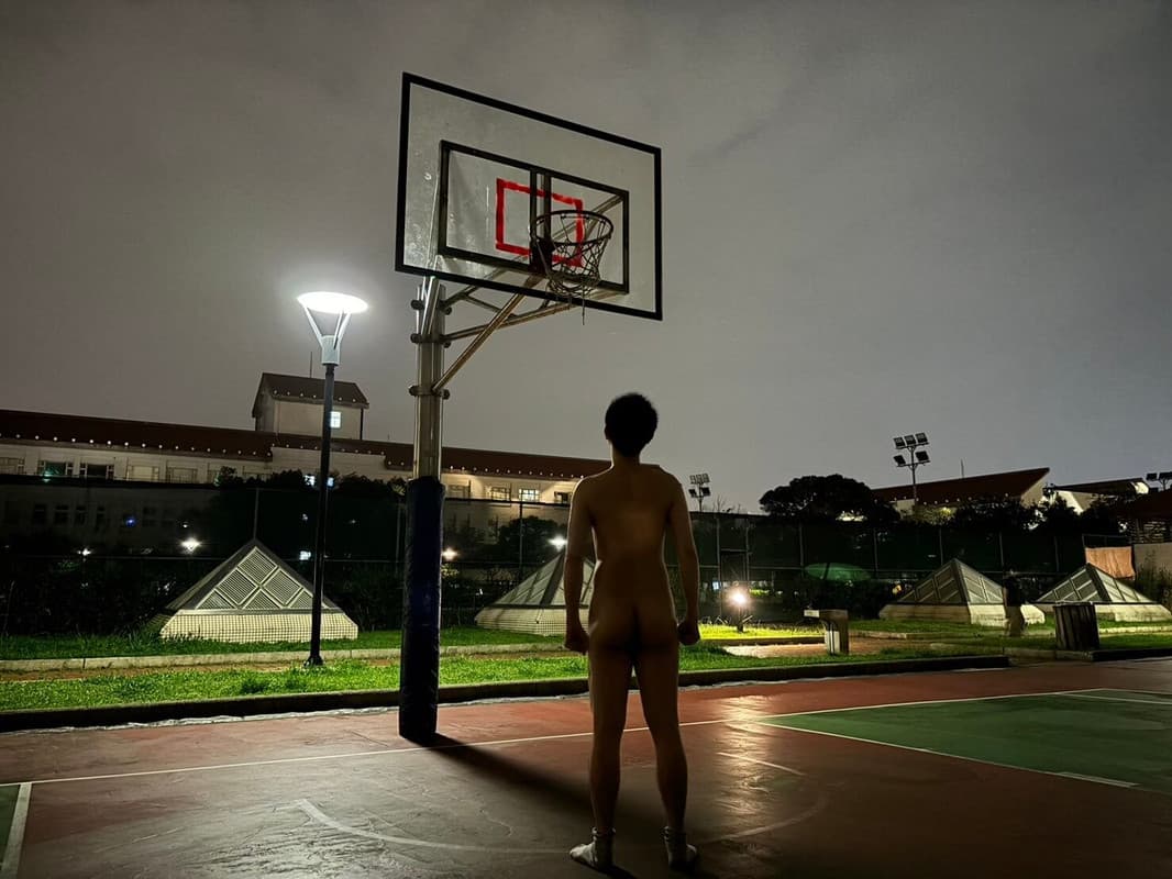 A nude figure stands alone on an empty basketball court at night, silhouetted under a streetlamp while gazing up at the hoop. The scene captures profound vulnerability and quiet introspection against the dark institutional backdrop.