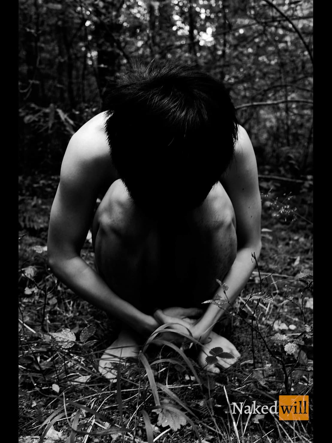A shirtless man with long dark hair sits curled in a fetal-like pose amid forest foliage, his face hidden in shadow as he hugs his knees, evoking isolation and quiet introspection through the stark black-and-white composition and dappled light filtering through the trees.