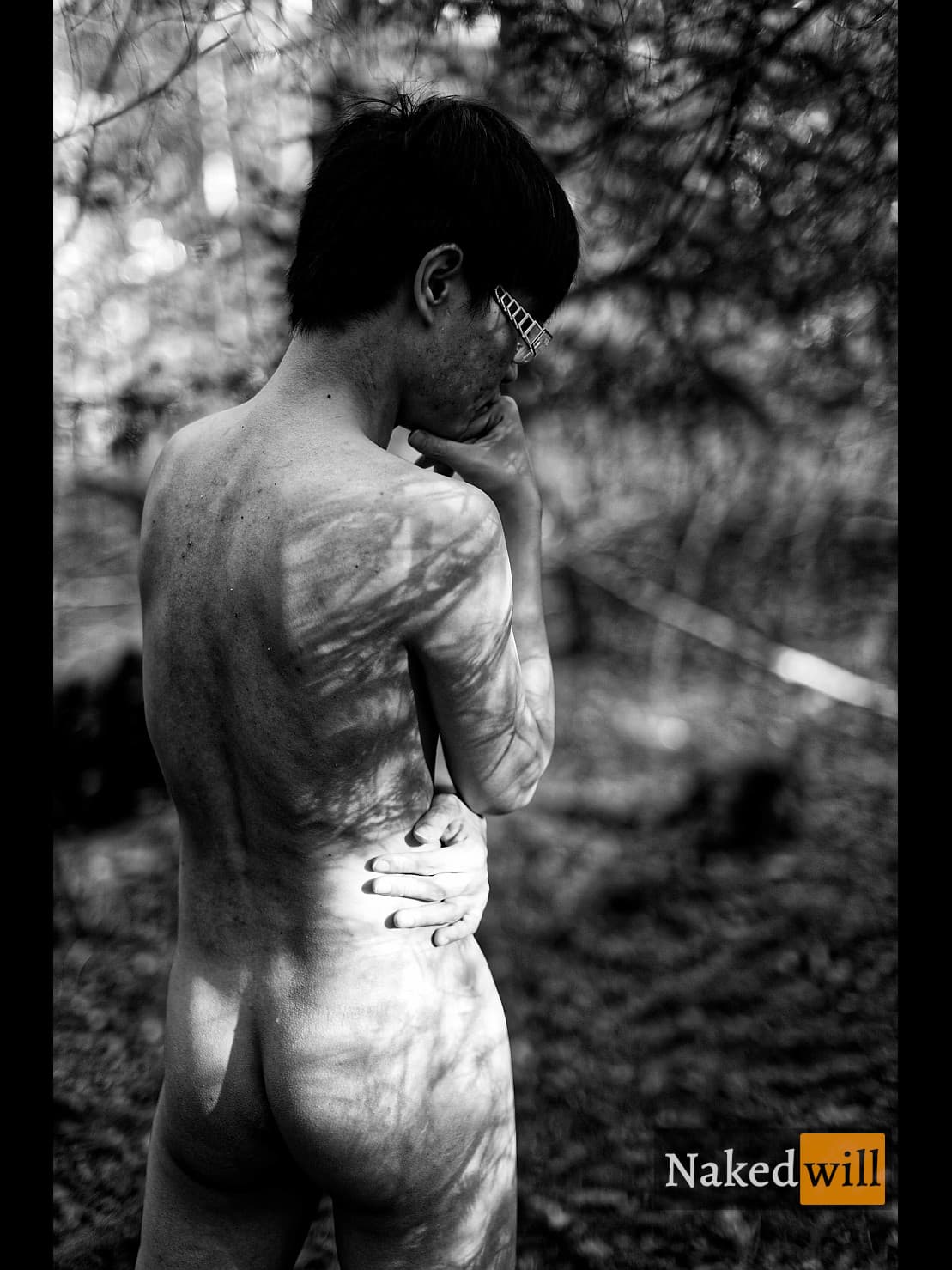 A nude young man stands pensively in a forest, one hand on his chin in deep thought as dappled sunlight casts intricate shadows across his bare back and form. The contemplative pose merges human vulnerability with nature's play of light and texture.